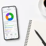Android smartphone displaying a budget tracking app with charts and graphs on a desk